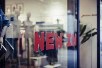 Red letters spelling 'NEW IN' are prominently displayed on a glass window, hinting at the entrance to a retail store. Inside, mannequins and display racks featuring clothing items can be seen, indicating a fashion shop setup. Blurred elements and reflections add depth to the scene.