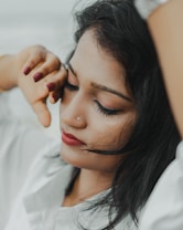 A close-up of a woman resting with her eyes closed. Her hand is gently touching her face, and she has a serene and peaceful expression. She is wearing a white shirt, and her dark hair is casually draped around her face. A small, delicate nose stud adds a touch of elegance.