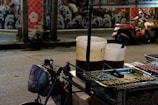 The image captures a night street food scene with a small vendor cart displaying skewers of various grilled foods. Two large plastic containers, likely holding sauces or soup, are placed on the cart. A scooter is parked on the street nearby, and the background features a mural with wave and flower patterns.