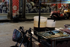 The image captures a night street food scene with a small vendor cart displaying skewers of various grilled foods. Two large plastic containers, likely holding sauces or soup, are placed on the cart. A scooter is parked on the street nearby, and the background features a mural with wave and flower patterns.