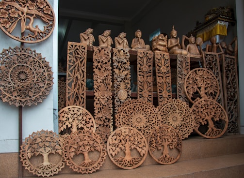 Intricately designed wooden carvings featuring circular patterns, trees, and floral motifs. Several Buddhist statues in various poses are arranged among the woodwork, creating a display of artistry and craftsmanship.