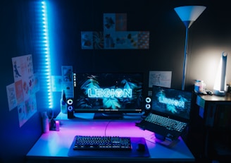 A modern computer setup with a large monitor and a laptop, both displaying the word 'LEGION' with a blue tropical leaf background. The desk is illuminated by blue and purple LED lights, with a mechanical keyboard, a gaming mouse, and speakers on either side of the monitor. The room is decorated with various posters on the wall. A floor lamp is situated on the right side, providing additional lighting.