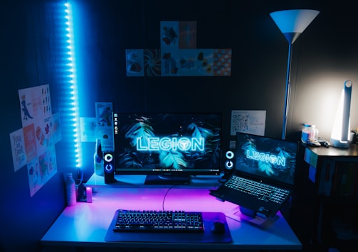 A modern computer setup with a large monitor and a laptop, both displaying the word 'LEGION' with a blue tropical leaf background. The desk is illuminated by blue and purple LED lights, with a mechanical keyboard, a gaming mouse, and speakers on either side of the monitor. The room is decorated with various posters on the wall. A floor lamp is situated on the right side, providing additional lighting.
