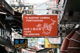 A busy urban street filled with various signs of shops and restaurants. The prominent sign in the center is for a Portuguese restaurant called 'O Santos' Comida Portuguesa located in Taipa, with contact information displayed. Below it, there is a sign for Koi Kei Bakery Macao written in both Chinese and English.