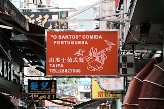 A busy urban street filled with various signs of shops and restaurants. The prominent sign in the center is for a Portuguese restaurant called 'O Santos' Comida Portuguesa located in Taipa, with contact information displayed. Below it, there is a sign for Koi Kei Bakery Macao written in both Chinese and English.