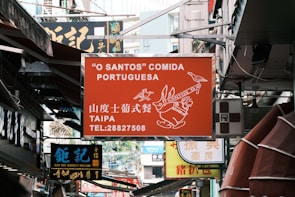 A busy urban street filled with various signs of shops and restaurants. The prominent sign in the center is for a Portuguese restaurant called 'O Santos' Comida Portuguesa located in Taipa, with contact information displayed. Below it, there is a sign for Koi Kei Bakery Macao written in both Chinese and English.