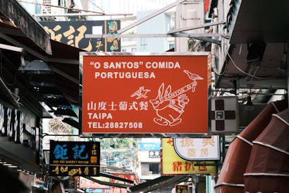 A busy urban street filled with various signs of shops and restaurants. The prominent sign in the center is for a Portuguese restaurant called 'O Santos' Comida Portuguesa located in Taipa, with contact information displayed. Below it, there is a sign for Koi Kei Bakery Macao written in both Chinese and English.