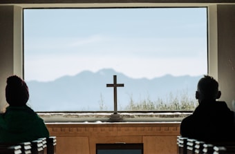 A serene interior setting featuring a large window overlooking a mountainous landscape. A cross is centered on the window sill, with two silhouetted figures on either side, likely seated on pews. The scene conveys tranquility and contemplation.