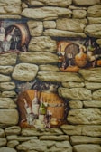 The image displays a rustic stone wall with several alcoves containing wine bottles, wooden barrels, glasses, and bunches of grapes. There are also baskets and assorted fruits such as apples, arranged artistically. The scene evokes the atmosphere of a wine cellar or vineyard setting.