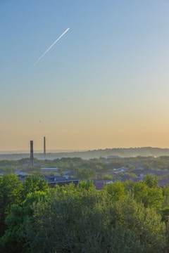A cityscape featuring industrial chimneys surrounded by dense greenery and tall trees. In the sky, a vapor trail from an airplane is seen against a gradient backdrop transitioning from blue to a warm yellow near the horizon. The distant horizon is lightly obscured by a haze.