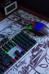 A desk setup featuring a RGB-backlit keyboard and a multicolored gaming mouse resting on a comic-themed mouse pad. An alarm clock displaying the time is positioned nearby.