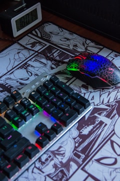 A desk setup featuring a RGB-backlit keyboard and a multicolored gaming mouse resting on a comic-themed mouse pad. An alarm clock displaying the time is positioned nearby.