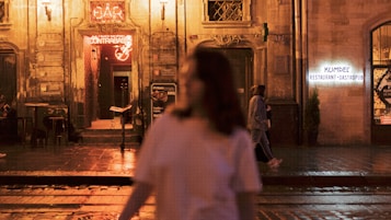 A nighttime street view featuring a warmly lit entrance to a bar with a neon sign above it. The background is an old stone building with atmospheric lighting. A person in a white shirt is blurred in the foreground, while two other people walk along the sidewalk. Another building hosts a sign for a restaurant and gastropub.