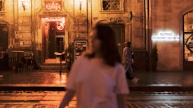 A nighttime street view featuring a warmly lit entrance to a bar with a neon sign above it. The background is an old stone building with atmospheric lighting. A person in a white shirt is blurred in the foreground, while two other people walk along the sidewalk. Another building hosts a sign for a restaurant and gastropub.