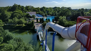A large waterslide descends into a pool surrounded by trees and green vegetation. The scene is part of a water park, with seating areas and paths visible in the background. The slide is a white, covered tube structure that ends in a splash zone near a tranquil pond.