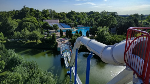 A large waterslide descends into a pool surrounded by trees and green vegetation. The scene is part of a water park, with seating areas and paths visible in the background. The slide is a white, covered tube structure that ends in a splash zone near a tranquil pond.