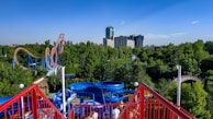 Amusement park scene featuring a vibrant blue and red waterslide in the foreground. A towering roller coaster with red tracks and blue supports snakes through lush green trees in the middle distance. In the background, modern high-rise buildings rise above the treetops under a clear blue sky.
