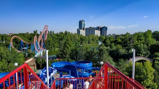 Amusement park scene featuring a vibrant blue and red waterslide in the foreground. A towering roller coaster with red tracks and blue supports snakes through lush green trees in the middle distance. In the background, modern high-rise buildings rise above the treetops under a clear blue sky.