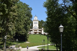 A grand villa situated atop a small hill is framed by lush green trees. Several pathways lead through the park area toward the villa, with vintage street lamps lining the paths. People are walking along the paths and enjoying the peaceful surroundings. The villa is an architectural highlight with a flat roof and multiple floors.