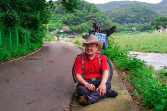 An elderly man wearing a red shirt and a straw hat is sitting on a paved path next to a river. He's smiling and seated near a wheelchair, surrounded by lush greenery. The path winds through the countryside, with trees and hills visible in the background.
