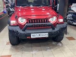 A red SUV with a rugged front bumper and circular LED headlights is parked indoors. The vehicle has a visible license plate and off-road tires, suggesting it is designed for rough terrains. The surrounding area appears to be a garage or workshop with various automotive parts and tools in the background.