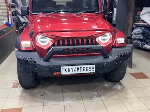 A red SUV with a rugged front bumper and circular LED headlights is parked indoors. The vehicle has a visible license plate and off-road tires, suggesting it is designed for rough terrains. The surrounding area appears to be a garage or workshop with various automotive parts and tools in the background.