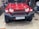 A red SUV with a rugged front bumper and circular LED headlights is parked indoors. The vehicle has a visible license plate and off-road tires, suggesting it is designed for rough terrains. The surrounding area appears to be a garage or workshop with various automotive parts and tools in the background.