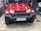 A red SUV with a rugged front bumper and circular LED headlights is parked indoors. The vehicle has a visible license plate and off-road tires, suggesting it is designed for rough terrains. The surrounding area appears to be a garage or workshop with various automotive parts and tools in the background.