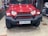 A red SUV with a rugged front bumper and circular LED headlights is parked indoors. The vehicle has a visible license plate and off-road tires, suggesting it is designed for rough terrains. The surrounding area appears to be a garage or workshop with various automotive parts and tools in the background.