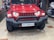 A red SUV with a rugged front bumper and circular LED headlights is parked indoors. The vehicle has a visible license plate and off-road tires, suggesting it is designed for rough terrains. The surrounding area appears to be a garage or workshop with various automotive parts and tools in the background.