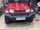 A red SUV with a rugged front bumper and circular LED headlights is parked indoors. The vehicle has a visible license plate and off-road tires, suggesting it is designed for rough terrains. The surrounding area appears to be a garage or workshop with various automotive parts and tools in the background.