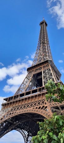 The image features the iconic Eiffel Tower against a bright blue sky, captured from a low angle. The intricate lattice structure of the tower is prominently displayed, and a tree with green leaves is visible in the foreground, adding a touch of nature to the scene.