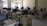 A classroom or computer lab setting with several workstations, each equipped with a computer monitor, keyboard, and mouse. The room has windows with bars that let in natural light, white tile flooring, and pale-colored walls. The desks are arranged in rows, and there are chairs in front of each workstation.