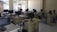 A classroom or computer lab setting with several workstations, each equipped with a computer monitor, keyboard, and mouse. The room has windows with bars that let in natural light, white tile flooring, and pale-colored walls. The desks are arranged in rows, and there are chairs in front of each workstation.
