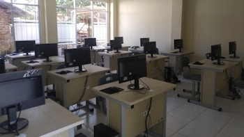 A classroom or computer lab setting with several workstations, each equipped with a computer monitor, keyboard, and mouse. The room has windows with bars that let in natural light, white tile flooring, and pale-colored walls. The desks are arranged in rows, and there are chairs in front of each workstation.