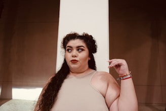 A person with long, dark, curly hair is wearing a sleeveless beige top and multiple bracelets on one arm. The background consists of a plain wall with contrasting light and shadow.