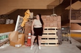 A person stands in an industrial or storage area, surrounded by building materials including wooden pallets, bricks, and a traffic cone. There is a shopping cart nearby and stacks of various construction materials like tiles and sacks are visible in the background.