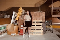 A person stands in an industrial or storage area, surrounded by building materials including wooden pallets, bricks, and a traffic cone. There is a shopping cart nearby and stacks of various construction materials like tiles and sacks are visible in the background.