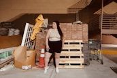 A person stands in an industrial or storage area, surrounded by building materials including wooden pallets, bricks, and a traffic cone. There is a shopping cart nearby and stacks of various construction materials like tiles and sacks are visible in the background.