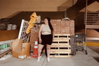 A person stands in an industrial or storage area, surrounded by building materials including wooden pallets, bricks, and a traffic cone. There is a shopping cart nearby and stacks of various construction materials like tiles and sacks are visible in the background.