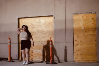 A person stands in front of a boarded-up doorway next to a construction barrier. The person's hand is raised to their head. The setting appears to be a construction site or an area undergoing renovation.