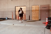 A person walks on a concrete platform adjacent to a boarded-up building. Wooden panels cover some of the entryways, and the door marked with 'stairwell' indicates an industrial setting. The scene includes construction cones and various plastic barriers nearby, enhancing the urban atmosphere.