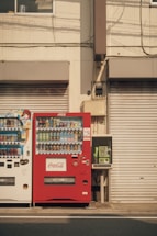 Two vending machines are positioned side by side against a beige building façade. The vending machine on the left is white and contains various drinks, while the one on the right is red and branded with Coca-Cola. To the right of the vending machines, there is a green public telephone. The shutters of the building are closed, and there are visible cables and pipes running along the wall.