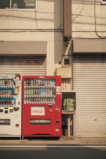 Two vending machines are positioned side by side against a beige building façade. The vending machine on the left is white and contains various drinks, while the one on the right is red and branded with Coca-Cola. To the right of the vending machines, there is a green public telephone. The shutters of the building are closed, and there are visible cables and pipes running along the wall.