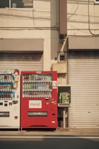 Two vending machines are positioned side by side against a beige building fa&ccedil;ade. The vending machine on the left is white and contains various drinks, while the one on the right is red and branded with Coca-Cola. To the right of the vending machines, there is a green public telephone. The shutters of the building are closed, and there are visible cables and pipes running along the wall.