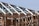 A partially constructed wooden framework of a building under a clear blue sky. The structure consists of exposed beams and trusses, showing the skeletal form of the roof and walls. Metal connectors are visible at the joints, enhancing the structural integrity.