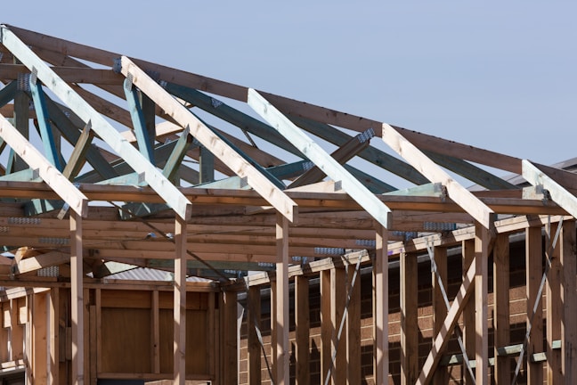 A partially constructed wooden framework of a building under a clear blue sky. The structure consists of exposed beams and trusses, showing the skeletal form of the roof and walls. Metal connectors are visible at the joints, enhancing the structural integrity.