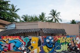 A vibrant mural covers a wall with various graffiti art including a depiction of a popular cartoon character with spiked hair and a blue monster with swirling eyes holding a green bottle. The background features palm trees and a partially visible building with a sloped roof.