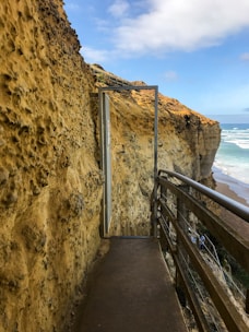 A narrow pathway carved into a rugged, sandy cliff leads to a steel-framed glass door. The pathway has a wooden railing on the right, overlooking a coastal view with waves crashing against the sand. The sky above is partly cloudy, enhancing the natural scenery.