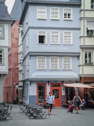 A charming cobblestone street scene featuring a three-story European building painted in pastel blue with white trim. The building has multiple windows, a small sign that reads 'Haus zur Flechte,' and a prominent red door. Adjacent outdoor patio seating with wrought iron tables and chairs is visible. Two people are casually strolling on the street, one pulling a red suitcase. The atmosphere is calm and leisurely, reminiscent of a quaint European town.